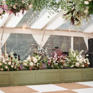 Elegant event stage decorated with lush floral arrangements and greenery, set in a tent for a wedding or special occasion, showcasing a wooden dance floor and musical instruments in the background.