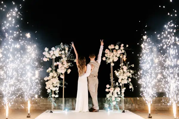 Young bride and groom stand near the wedding arch at night with lights, smoke and fireworks