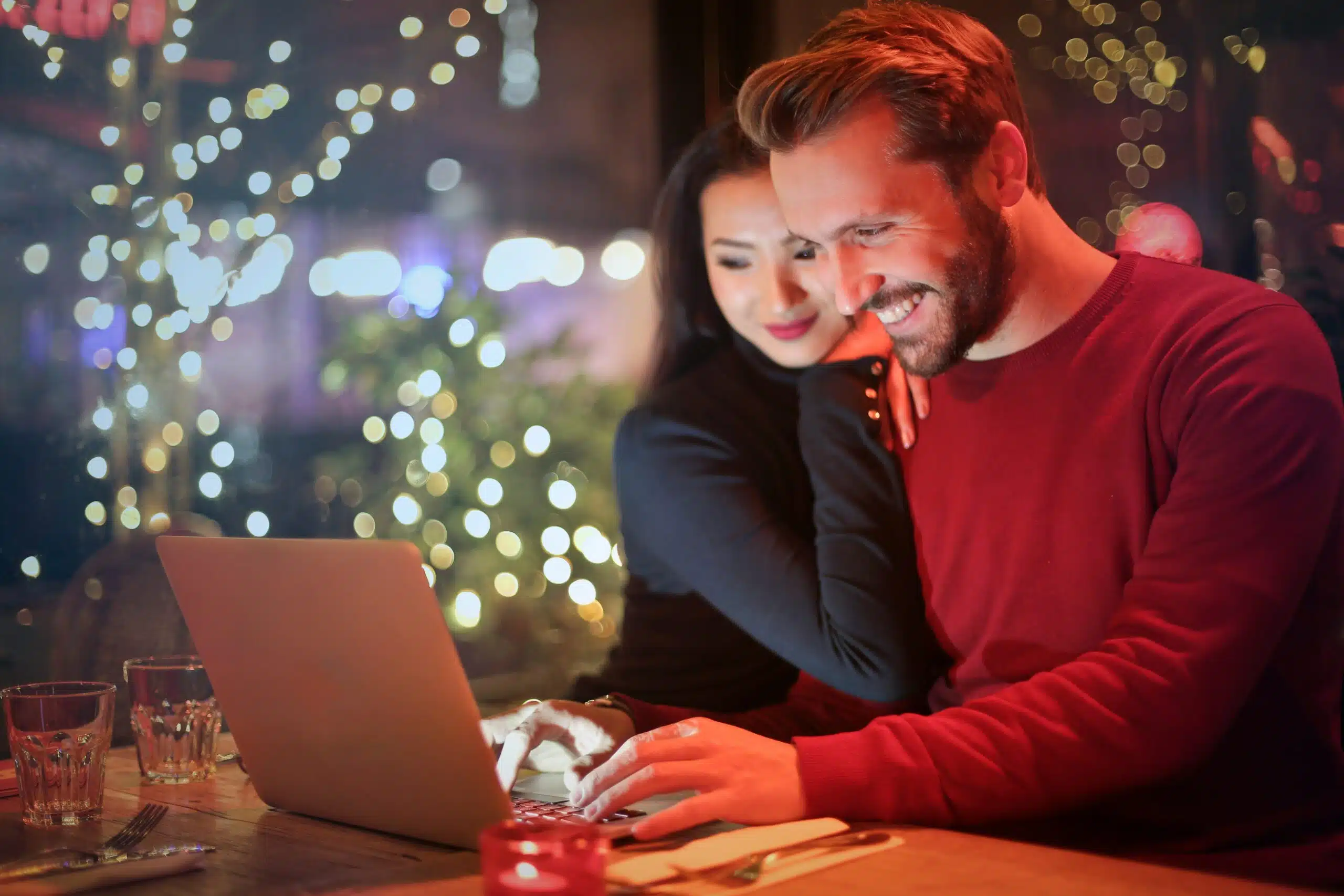 A smiling couple browses online together in a warmly lit setting, taking practical steps to incorporate themed furniture rentals into their upcoming celebration.