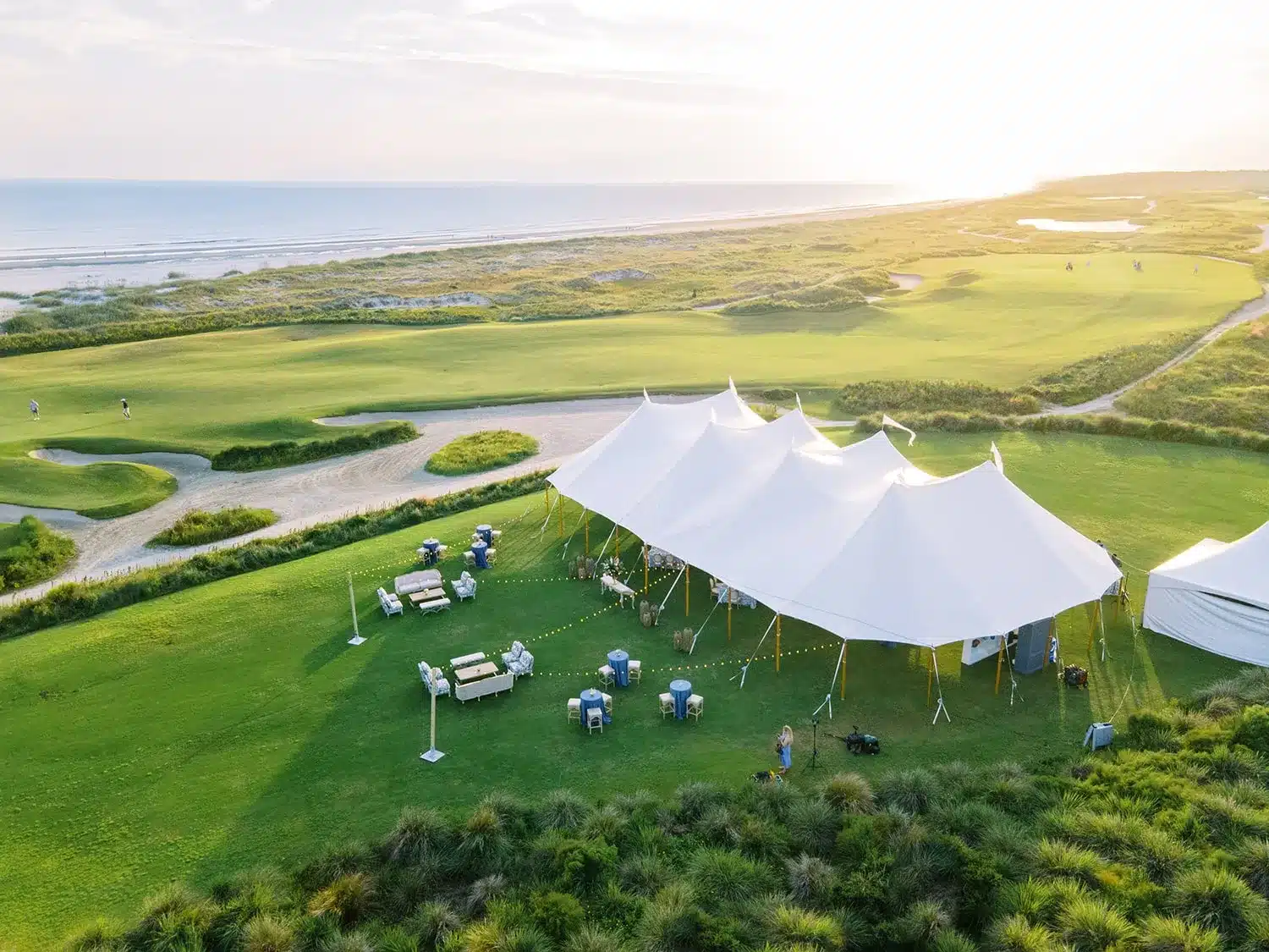Aerial view of a white sailcloth tent set on coastal green space for a beachfront event. | Curated Events Aerial view of a white sailcloth tent set on coastal green space for a beachfront event.