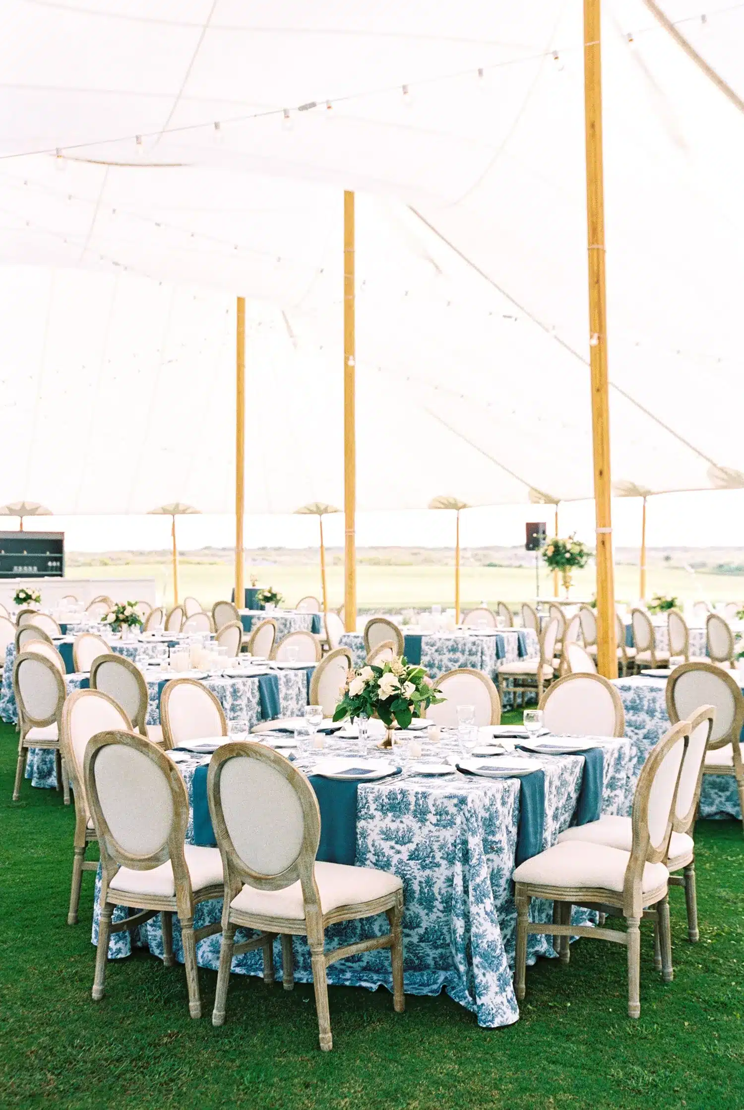 Upscale dining setup with toile tablecloths and round chairs under a sailcloth tent. | Curated Events Upscale dining setup with toile tablecloths and round chairs under a sailcloth tent.