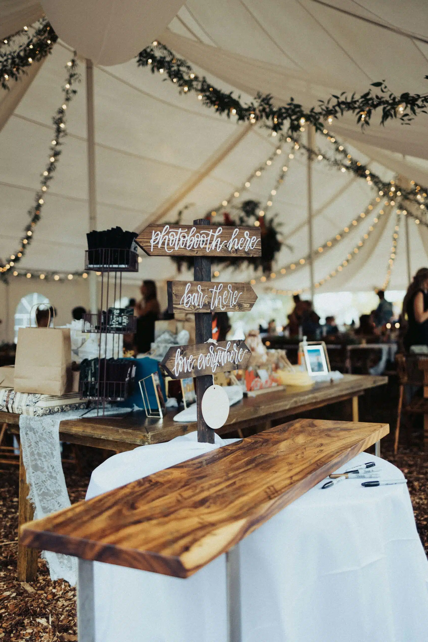 A wooden directional sign pointing to the bar and photobooth sits beside rustic décor under a canopy, highlighting a wedding bar setup that reflects the couple’s personal style and theme. | Curated Events A wooden directional sign pointing to the bar and photobooth sits beside rustic décor under a canopy, highlighting a wedding bar setup that reflects the couple’s personal style and theme.