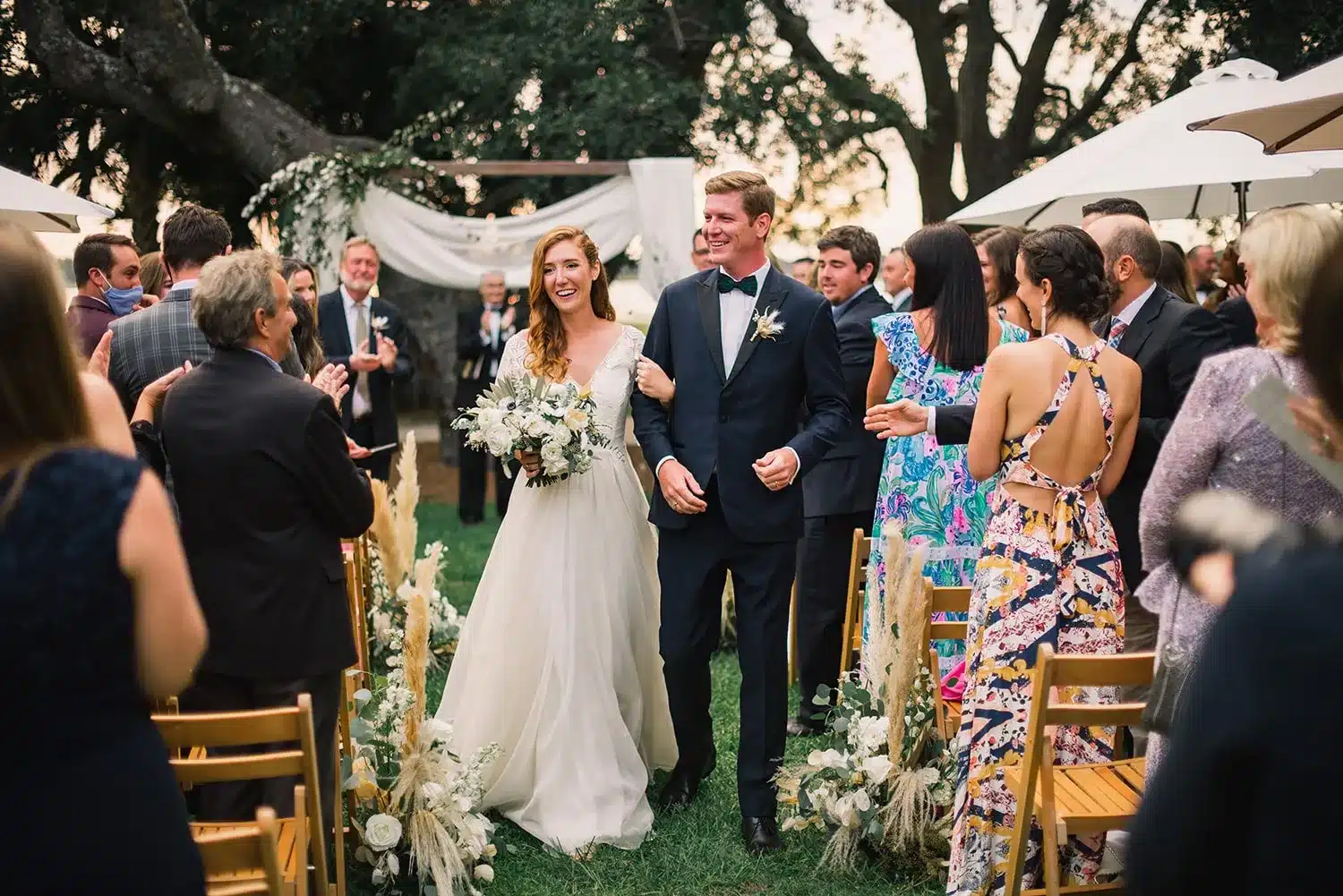 Bride and groom walk up the aisle smiling as guests cheer during a stylish outdoor Charleston wedding ceremony.