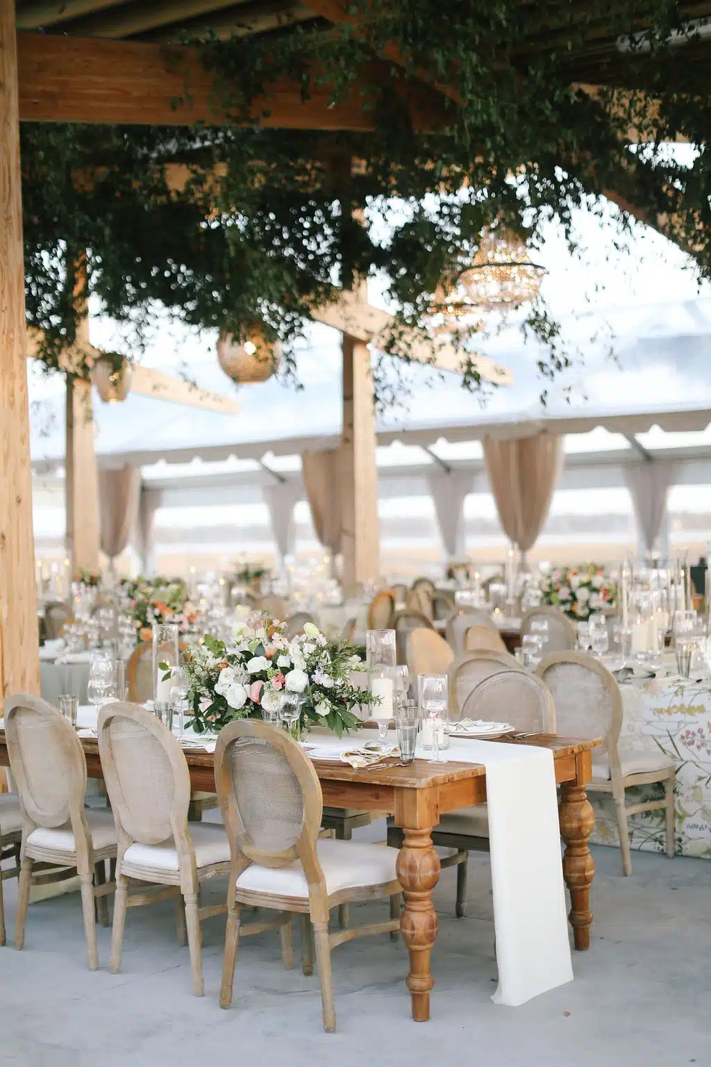 Elegant tented wedding reception with rustic wooden tables, floral centerpieces, and hanging greenery.