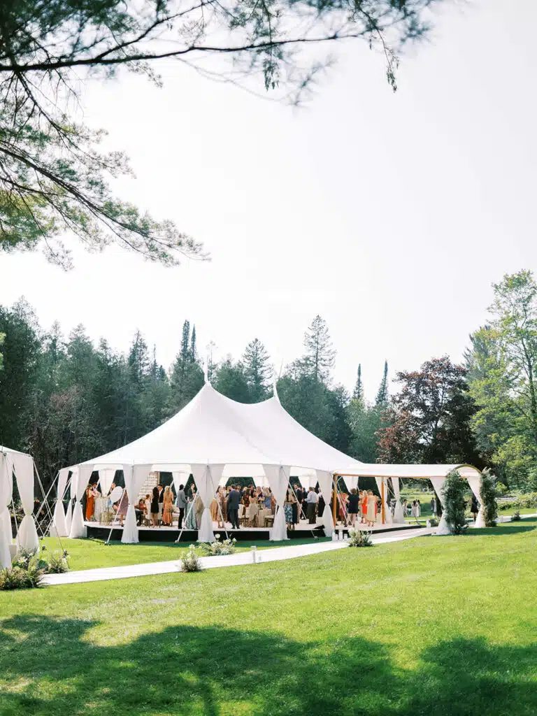 White peak-top tent surrounded by greenery with guests gathered inside for an outdoor wedding celebration.
