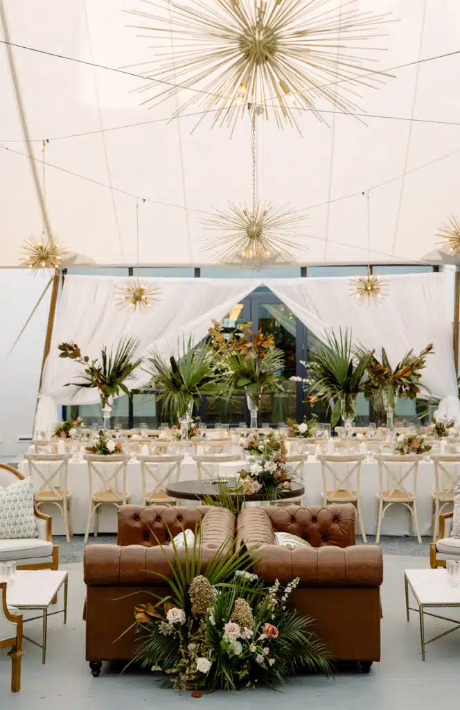 Luxury event lounge with tufted leather sofa, tropical centerpieces, and modern gold pendant lighting.
