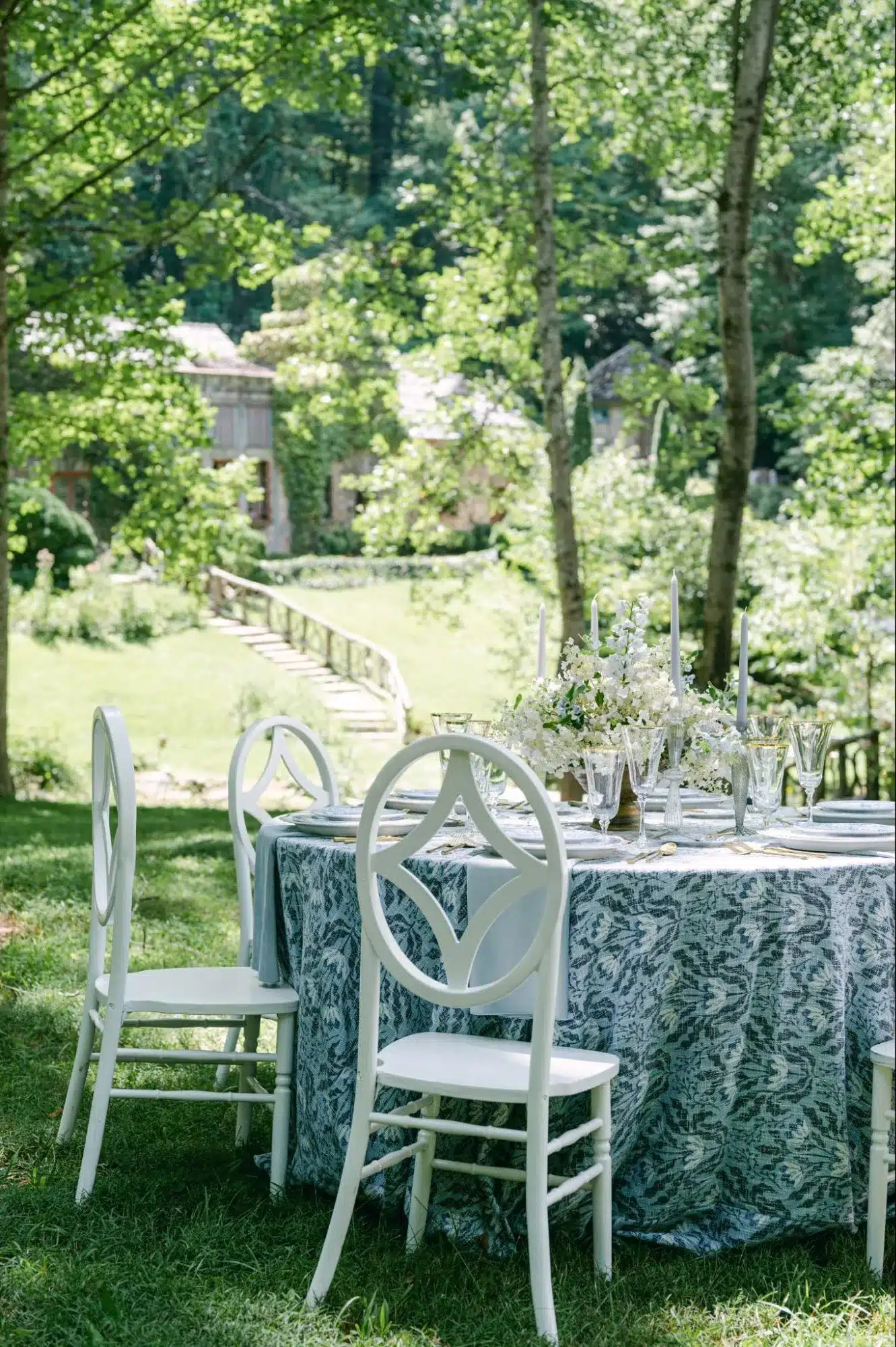 Round outdoor table with white chairs and blue patterned linen, styled with white florals and taper candles