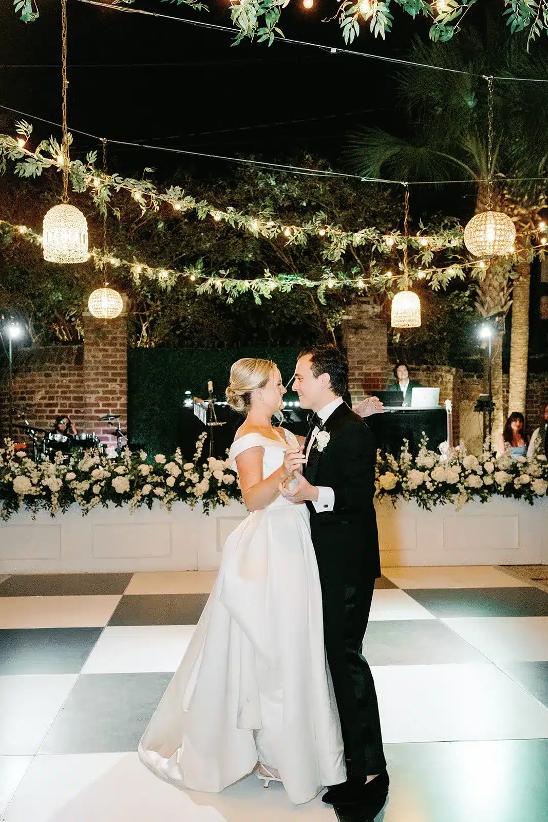 Bride and groom dancing on a black-and-white checkered dance floor under string lighting. | Curated Events Bride and groom dancing on a black-and-white checkered dance floor under string lighting.