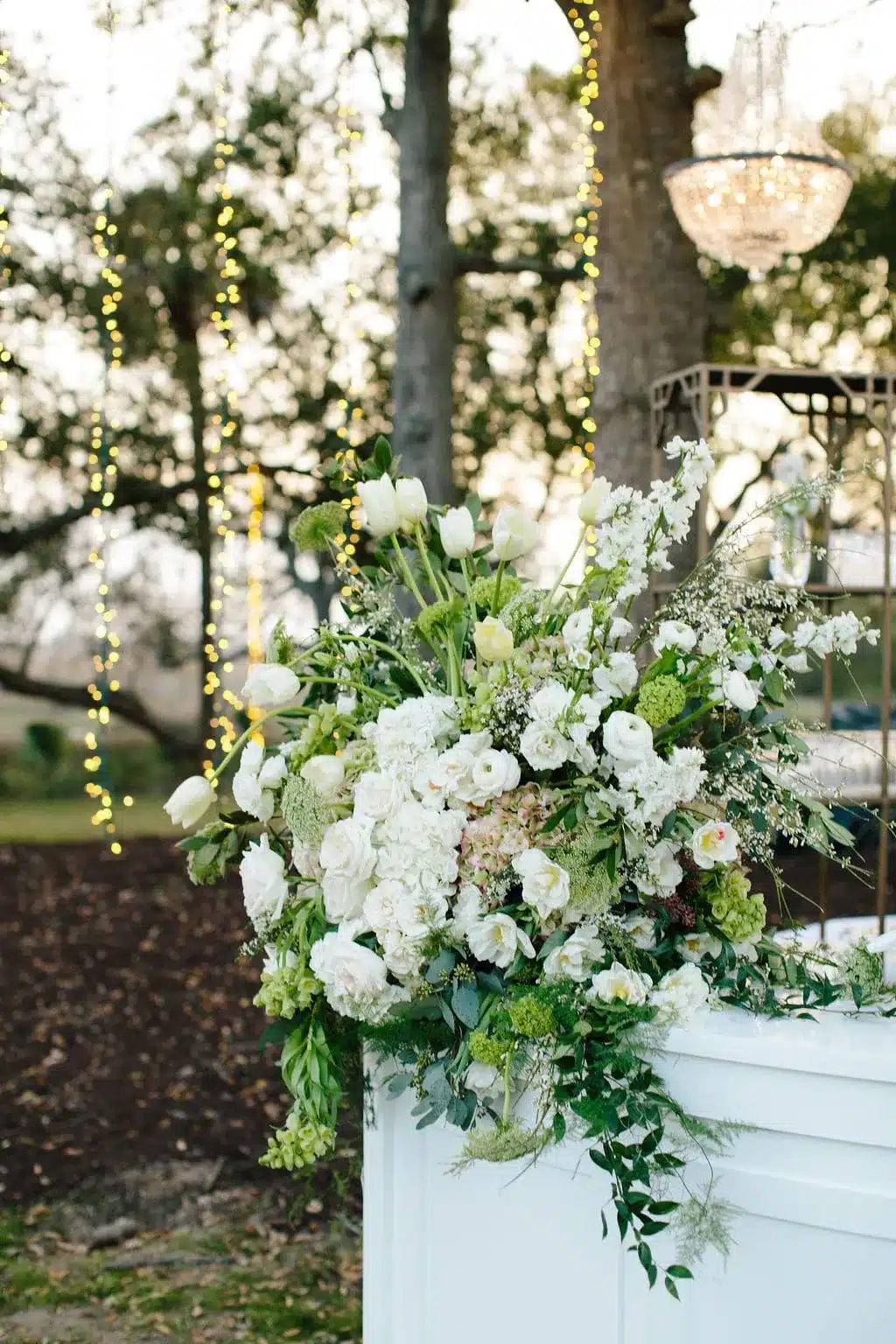Elegant white floral arrangement with tulips and roses beneath twinkling lights in an outdoor garden.