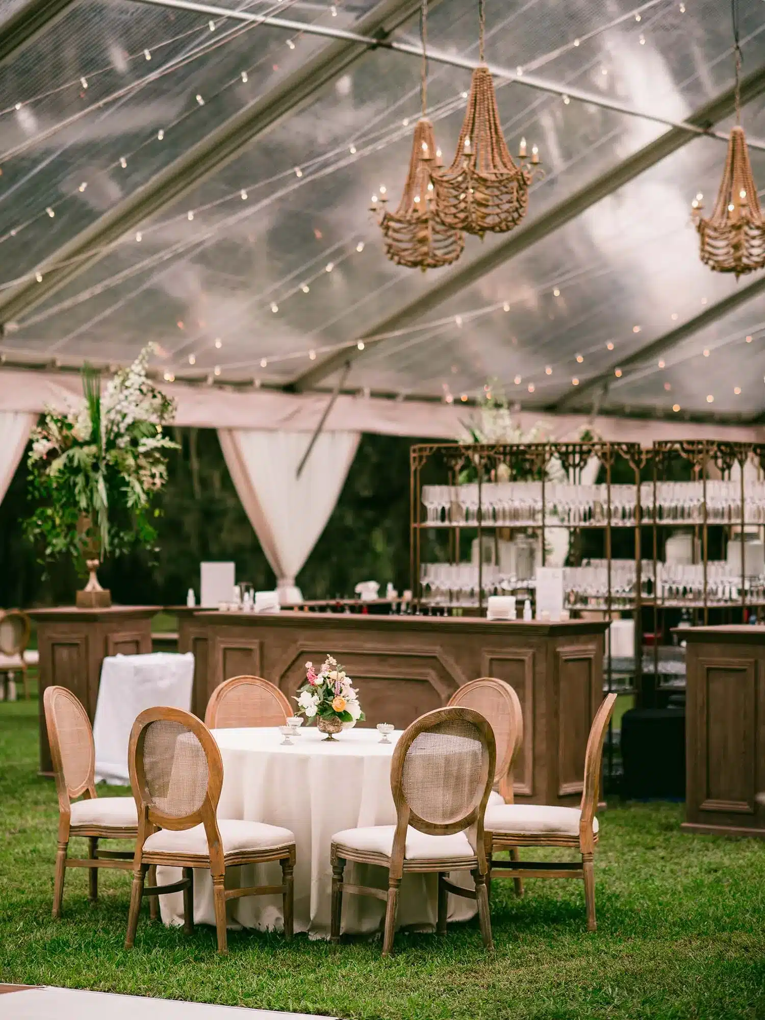 Elegant clear-top tent with wooden furniture, chandeliers, and lounge seating on grass