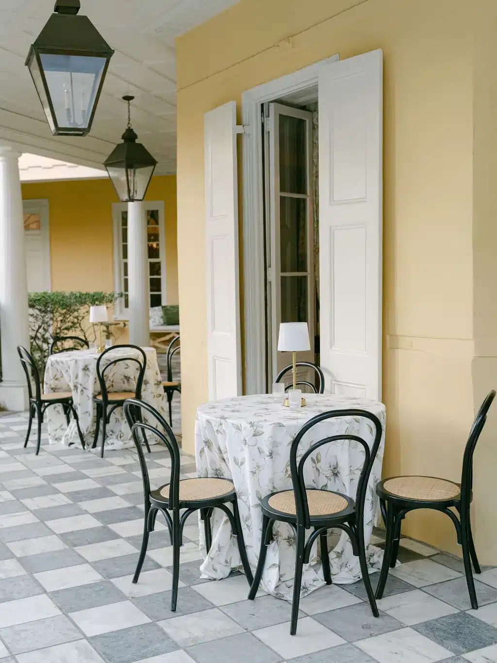 Outdoor café tables with black bistro chairs and floral tablecloths on checkered patio. | Curated Events Outdoor café tables with black bistro chairs and floral tablecloths on checkered patio.