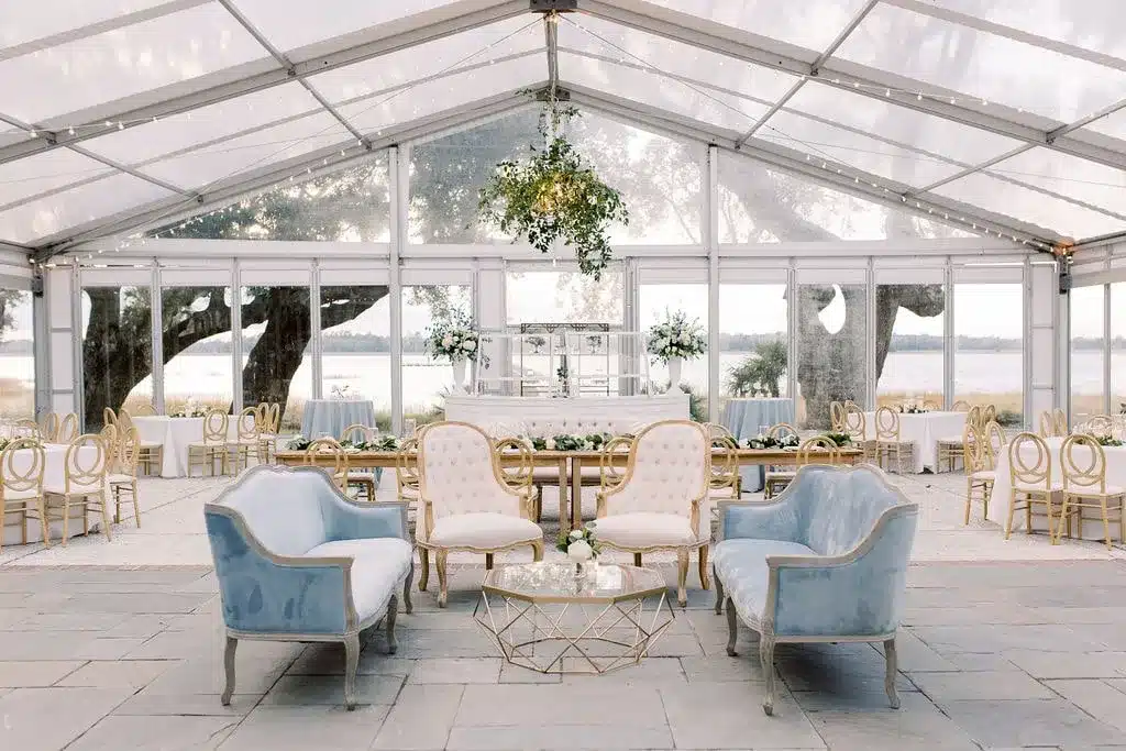 Micro-wedding lounge area under a clear tent with soft blue velvet and cream tufted furniture.
