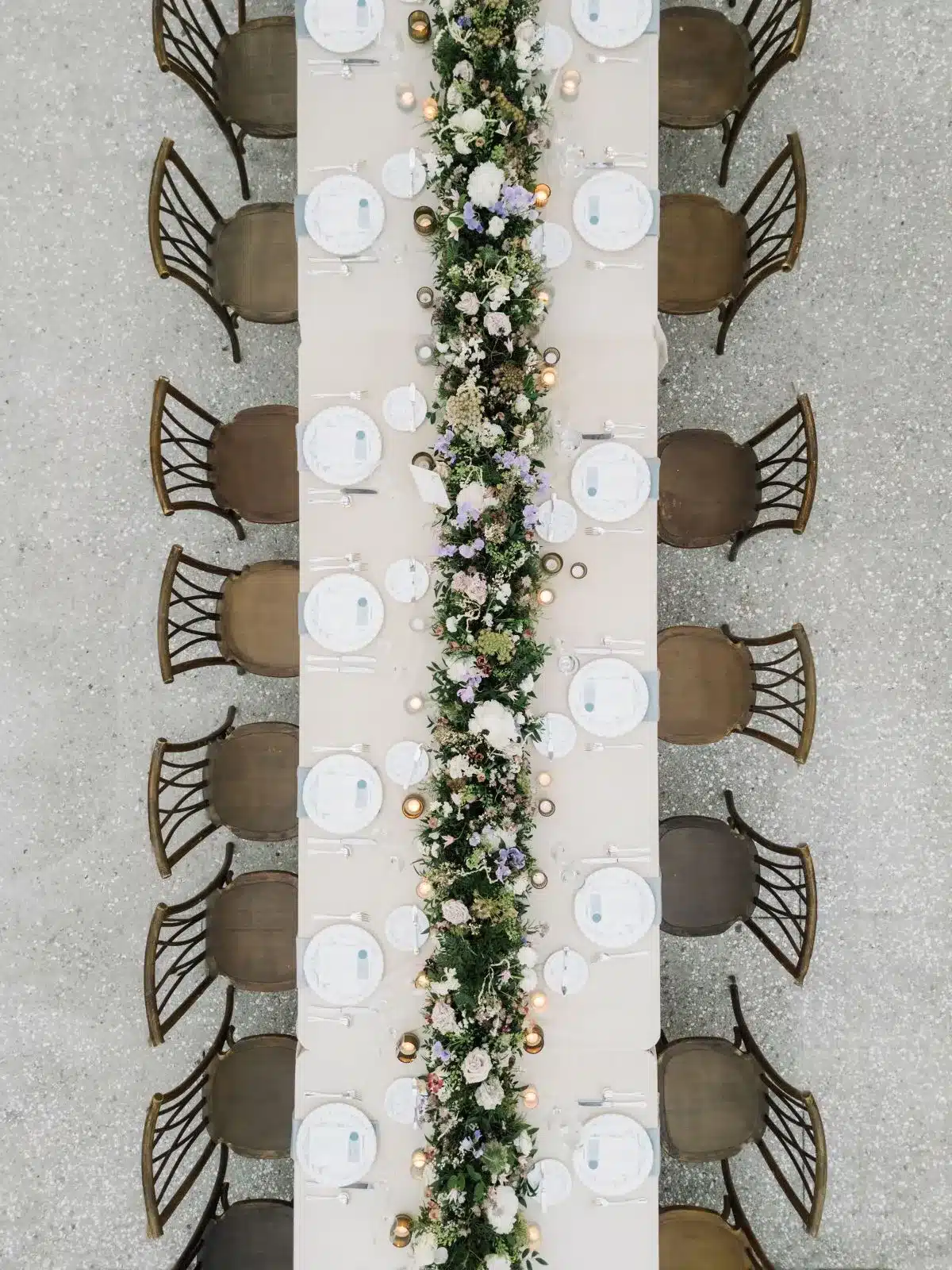 Overhead view of a long rectangular table with a lush floral garland centerpiece and wooden chairs.
