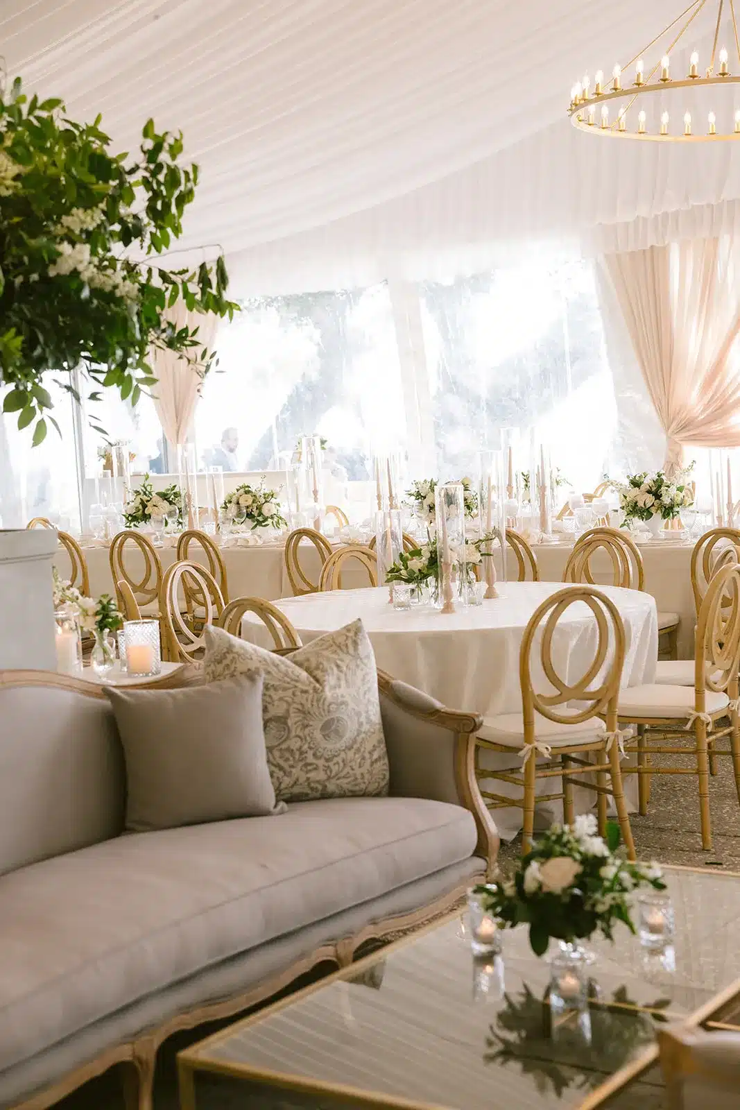 Interior of a clear event tent featuring ivory lounge seating and round tables with floral centerpieces.