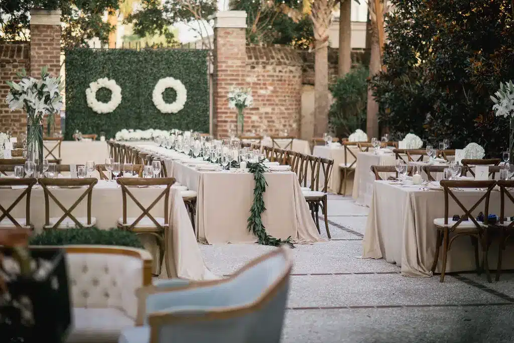 Modern outdoor event setup with long beige tables, wooden chairs, and boxwood backdrop.