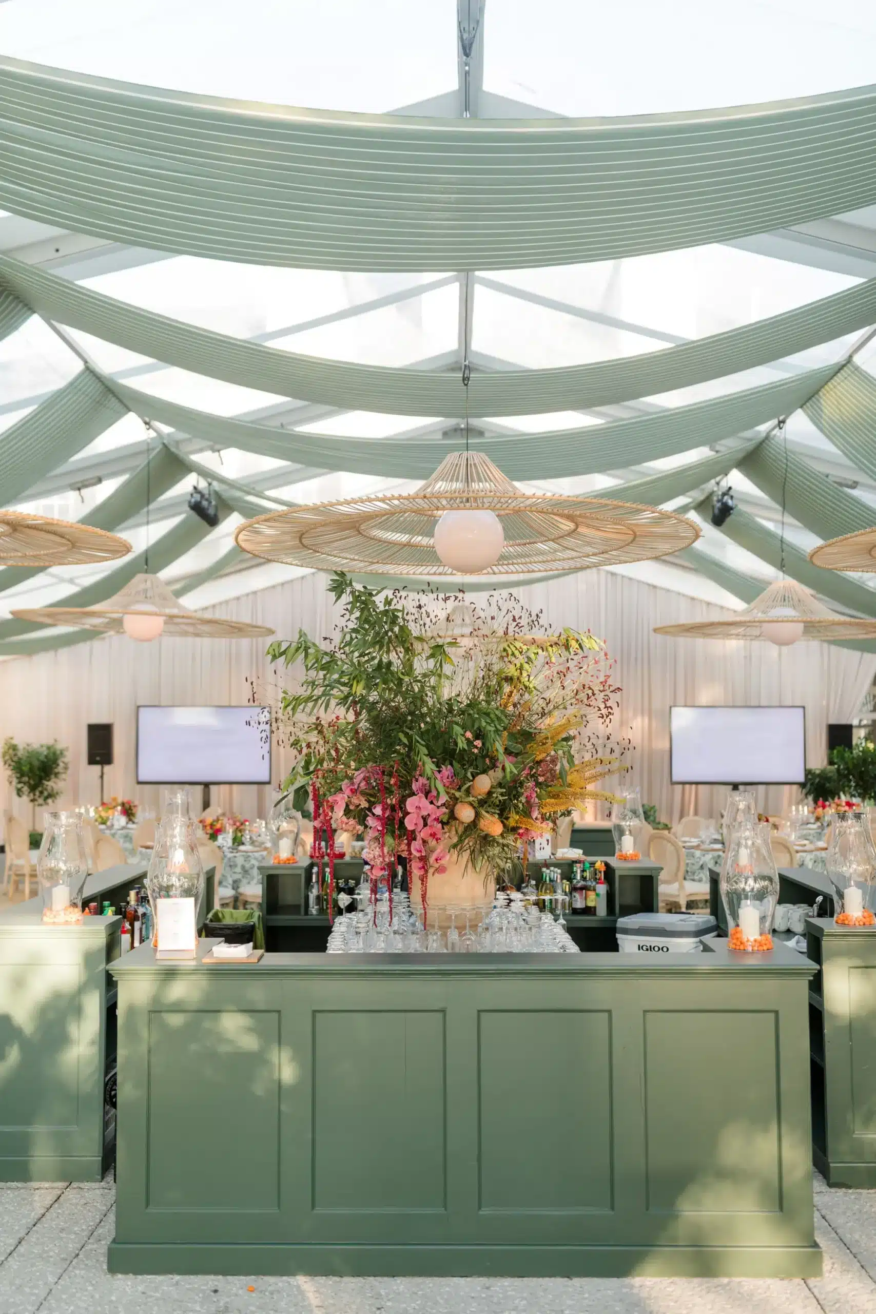 Stylish green bar with floral arrangement under a clear tent with draped ceiling and woven pendants.