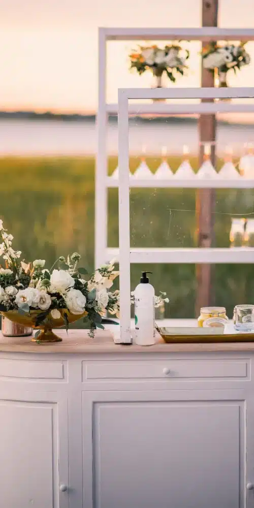 Outdoor bar setup with white cabinetry, floral arrangements, and glassware at sunset.