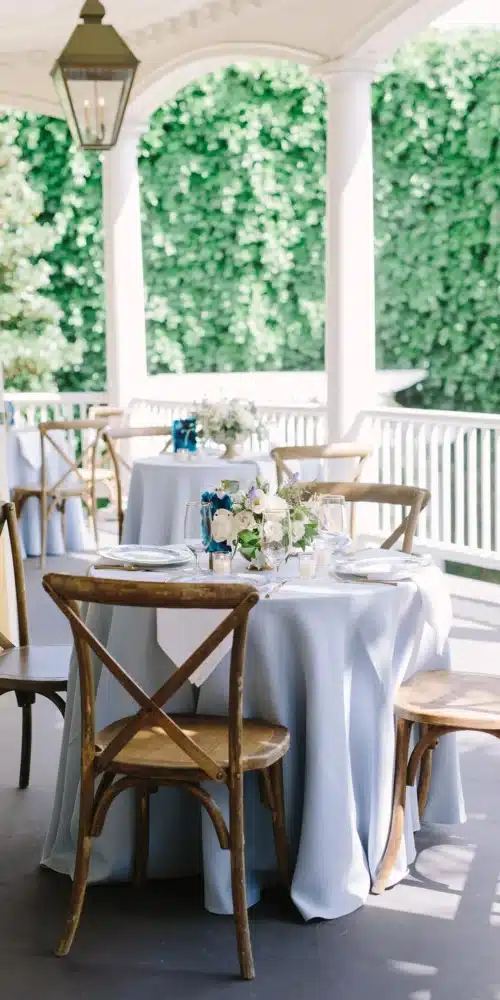 Outdoor rehearsal dinner table setup with blue linens, floral centerpieces, and wooden chairs on a veranda.
