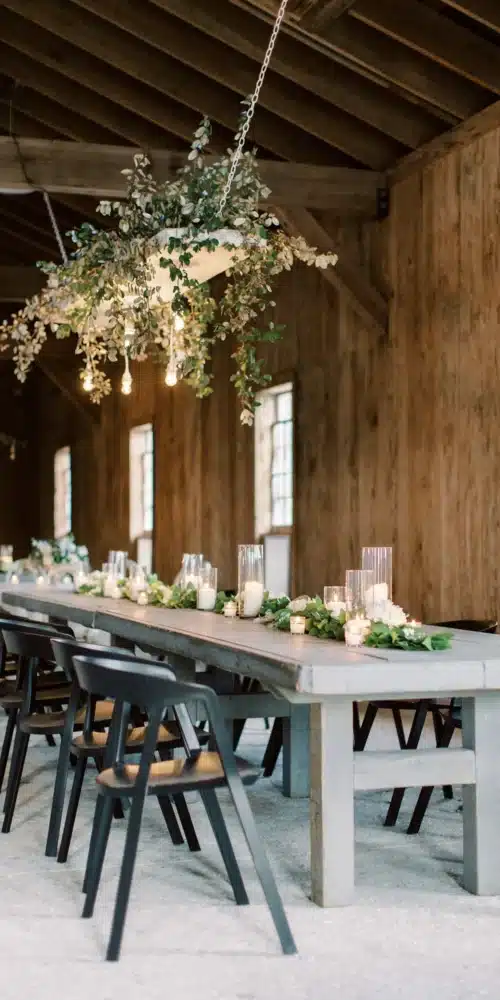 Candlelit tables with greenery and hanging floral d&eacute;cor inside a rustic event space.