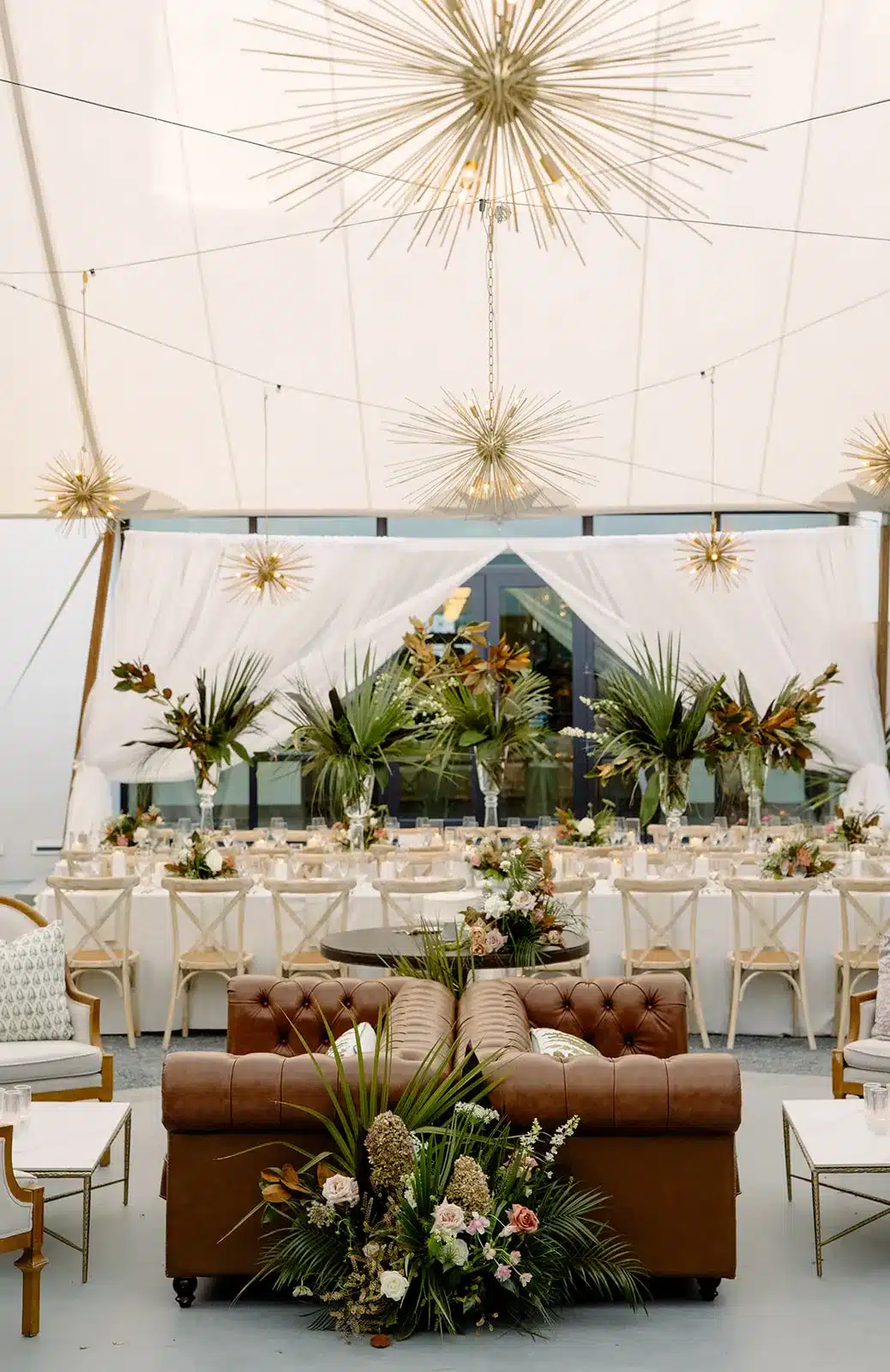 Elegant holiday tent interior with gold starburst chandeliers, lush greenery, and a refined dining setup.
