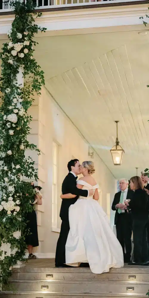 Bride and groom sharing a kiss on a flower-adorned porch.