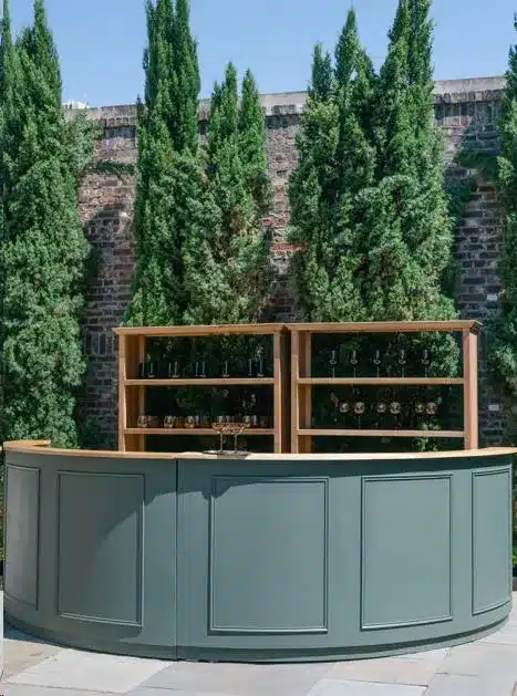 Curved sage-green bar front with shelving and glassware against a hedge wall.