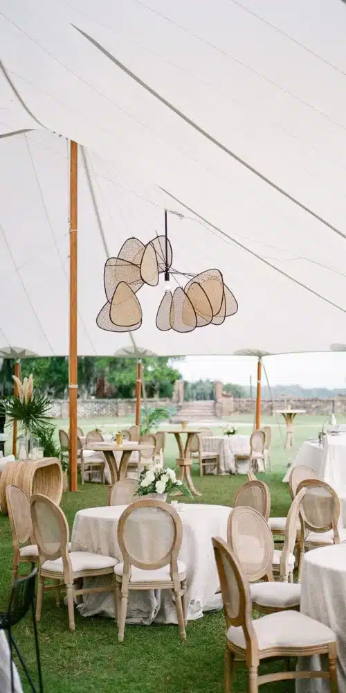 Corporate gala seating under a sailcloth tent with round tables, cane chairs, and sculptural pendant lighting.
