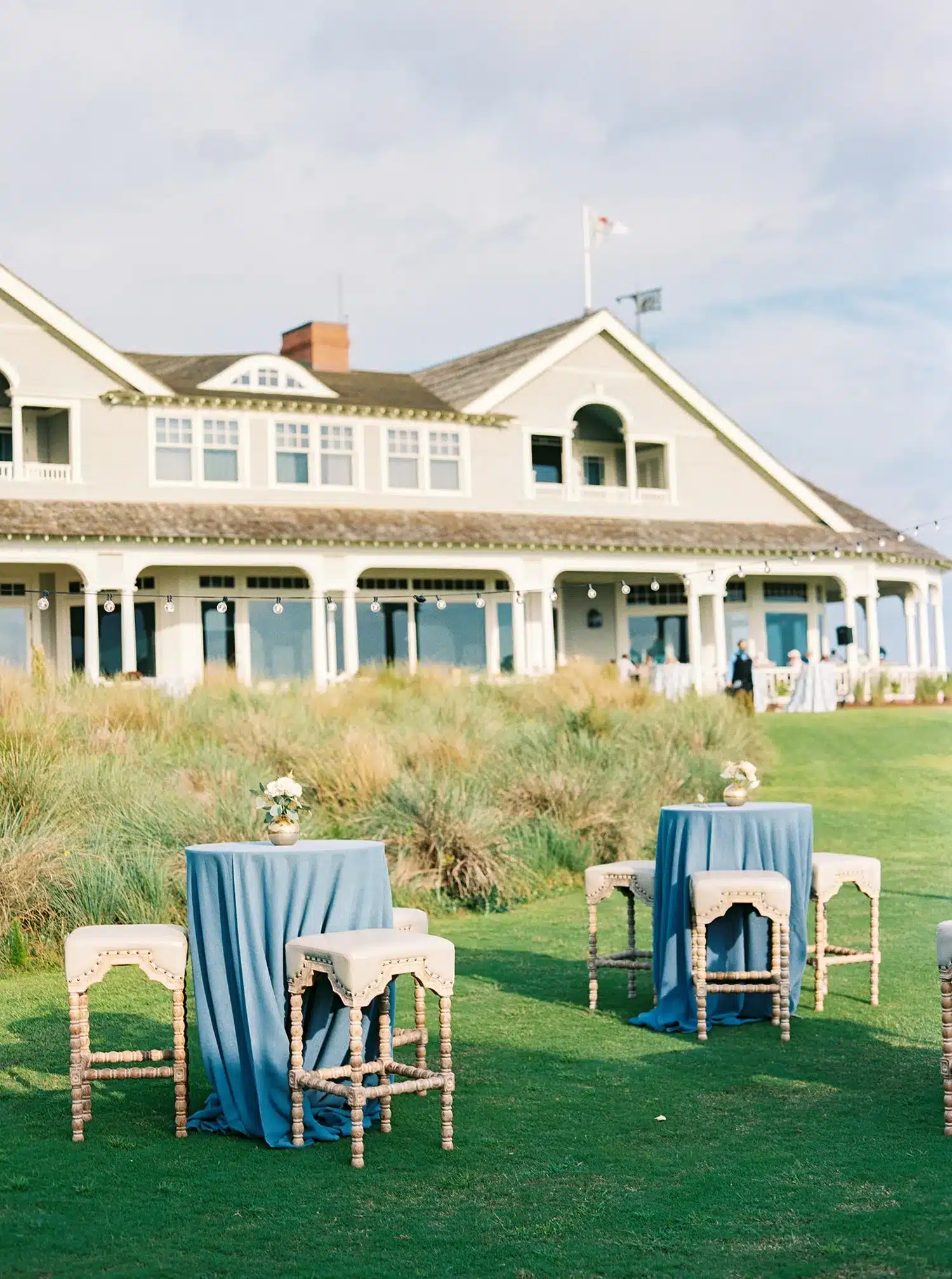 Coastal lawn cocktail setup with blue linens and stools in front of a waterfront clubhouse.