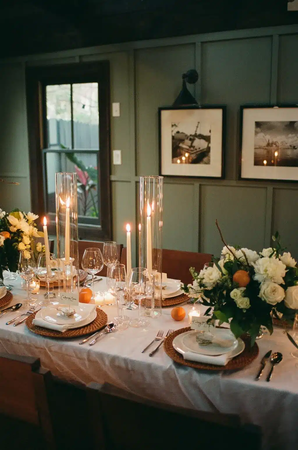 Candlelit indoor dining table with white florals, tall glass cylinders, and place settings in a moody room.
