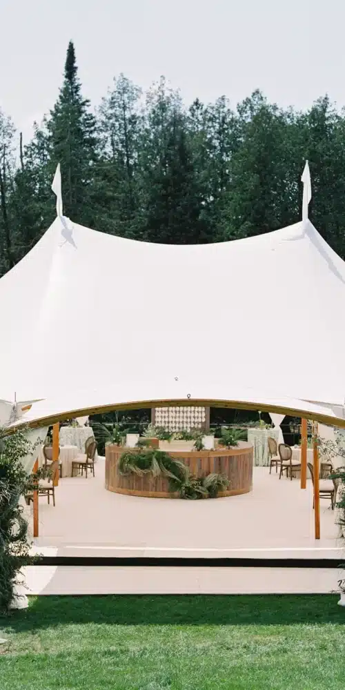 White sailcloth wedding tent with bar and seating in a wooded outdoor setting.