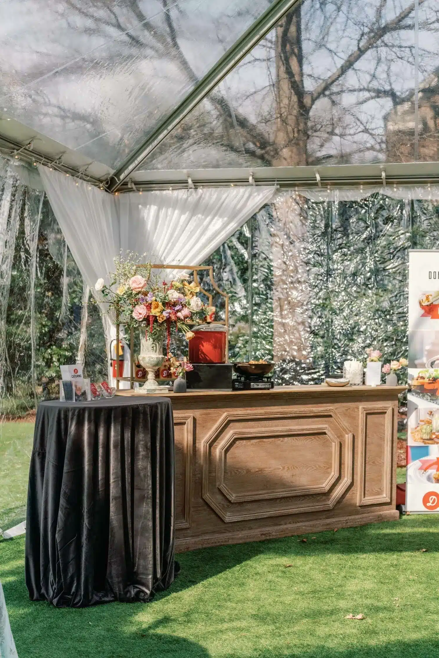 Wooden bar setup inside a clear-sided tent with draped curtains and floral decor.