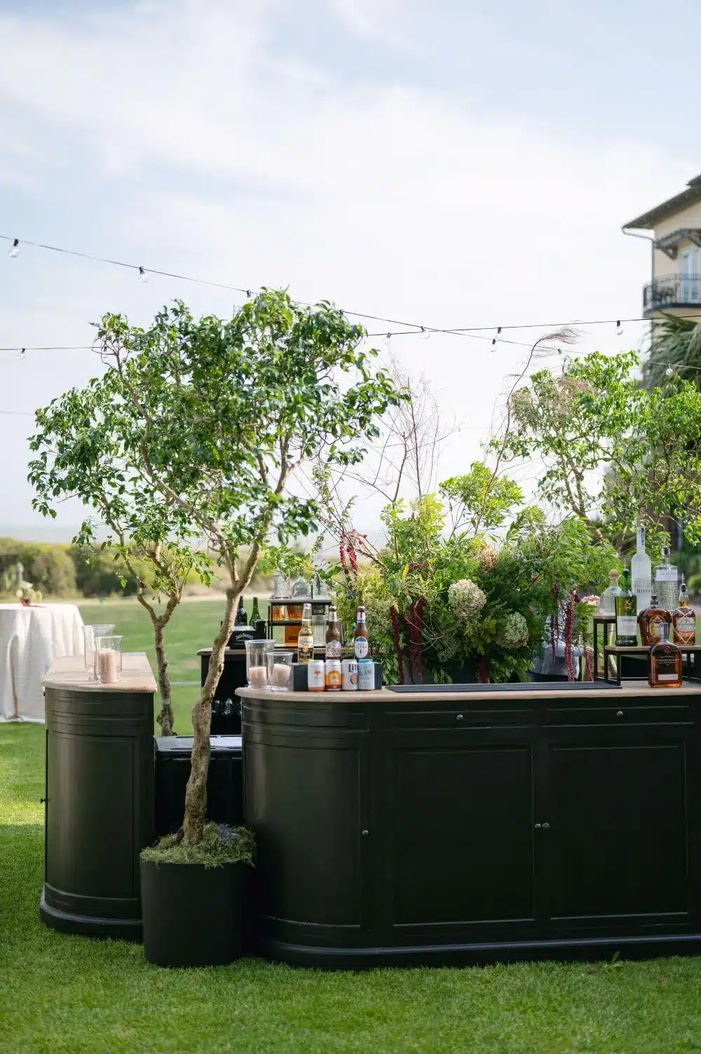 Outdoor wedding bar with greenery, string lights, and bottled spirits on display.