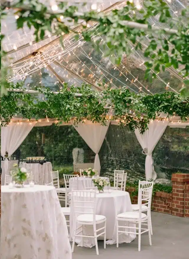 Clear-top tent with greenery garlands and white cocktail tables by a water feature.