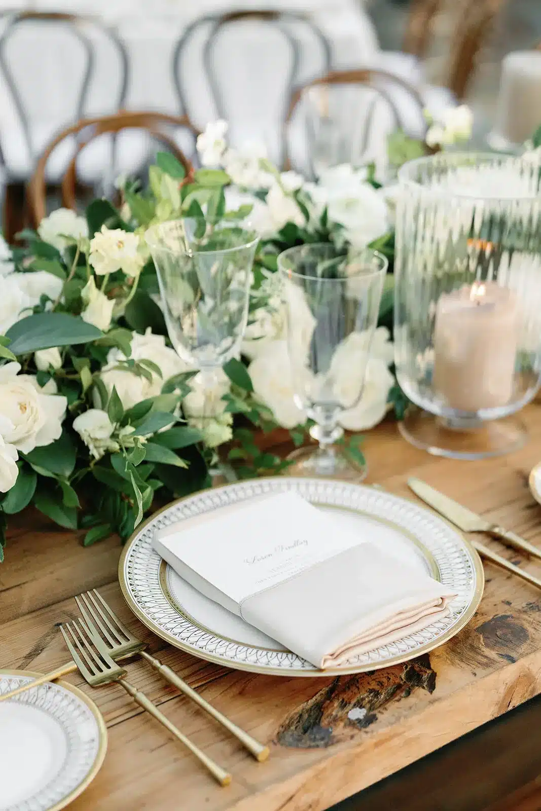 Elegant place setting with gold-rimmed china, neutral napkin, and lush greenery runner.