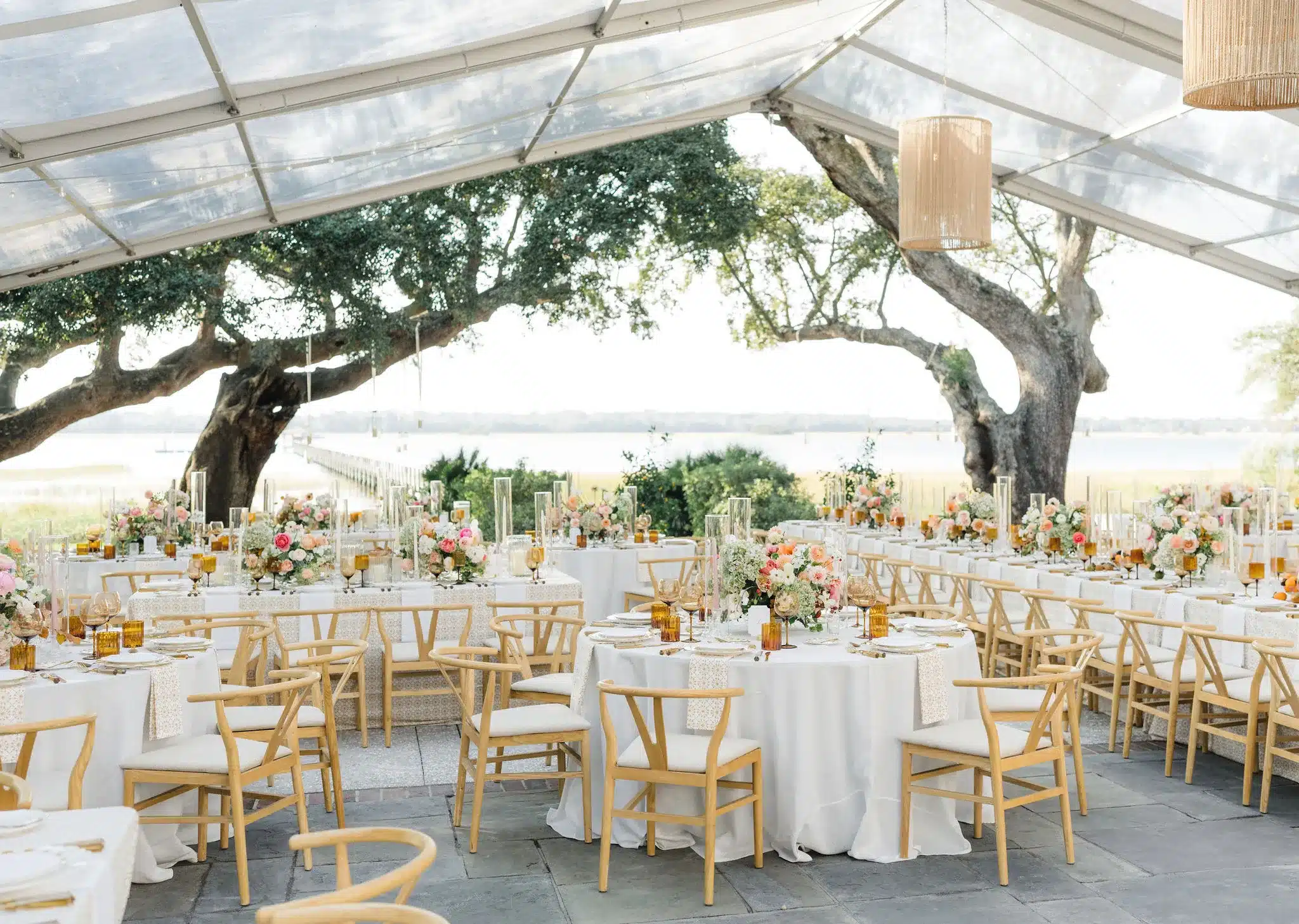 Clear-top tent reception with round tables and woven pendant lighting by the water.