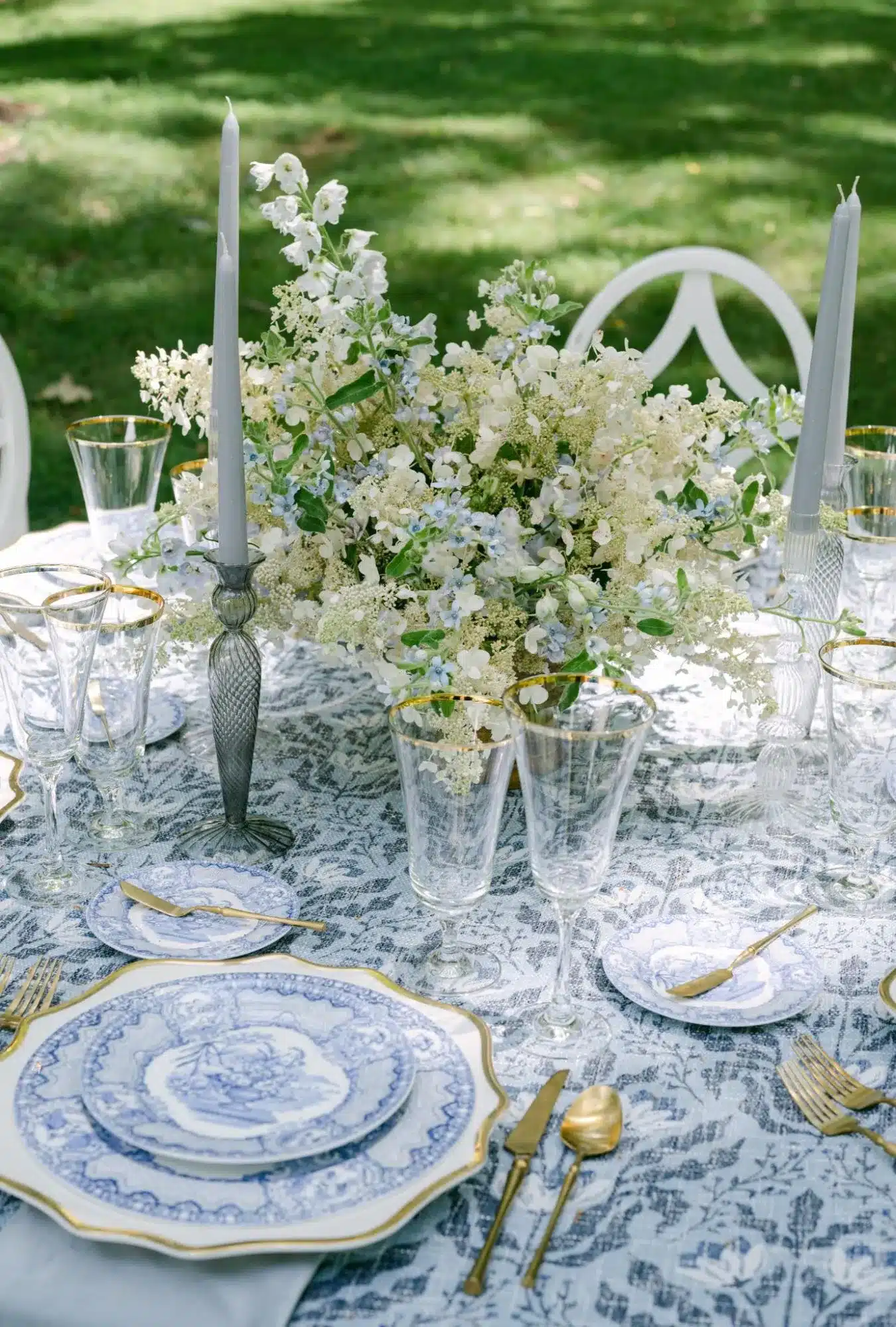 Elegant spring garden party table setting with blue and white china, gold-accented flatware, floral centerpiece, and glassware on a patterned tablecloth, surrounded by greenery.