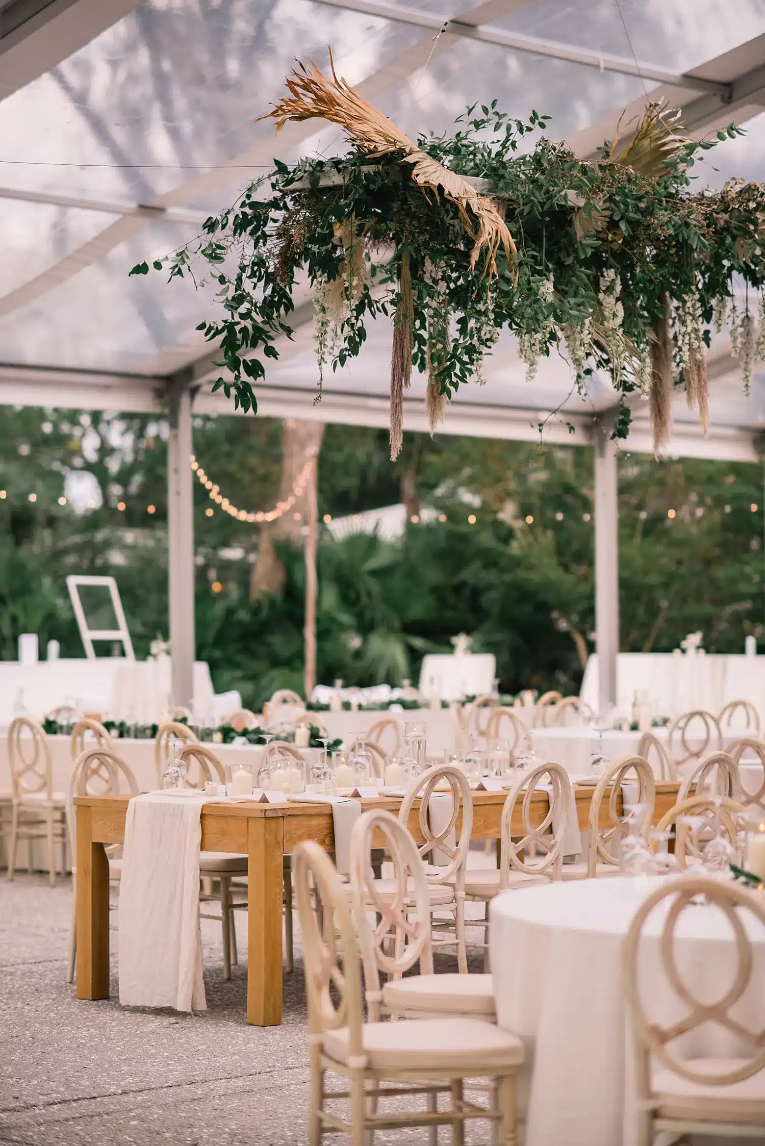 Tent reception with hanging greenery installation above wood and round dining tables.