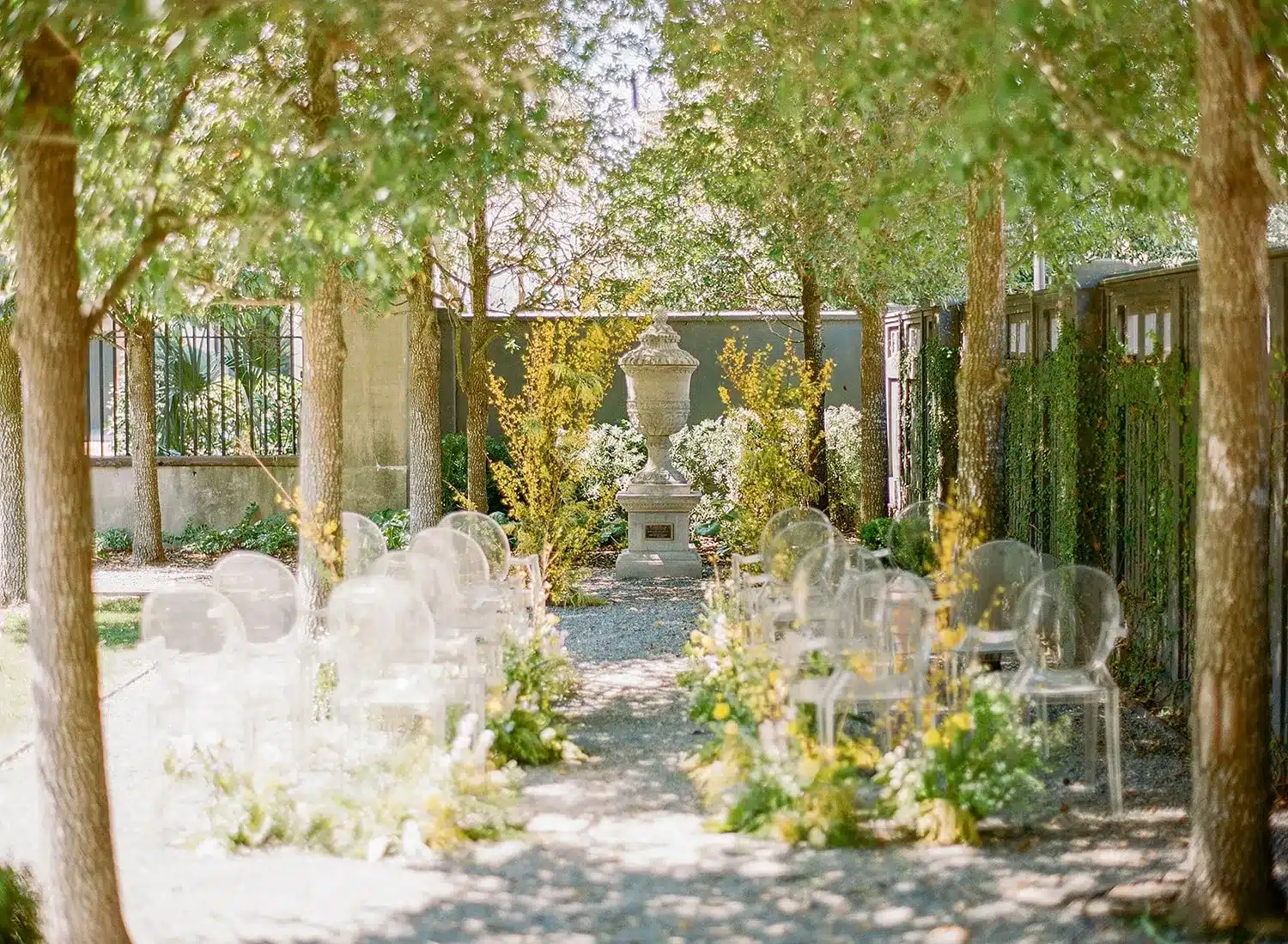 Clear acrylic chairs arranged along a pathway in a garden setting, flanked by greenery and yellow flowers, leading to a central stone urn, emphasizing accessibility and comfort for outdoor events.