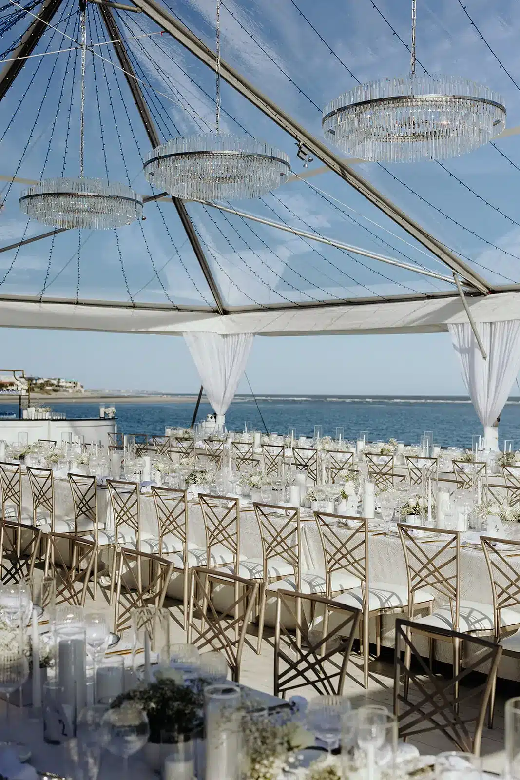 Oceanfront tented reception with long banquet tables, chandeliers, and cross-back chairs.