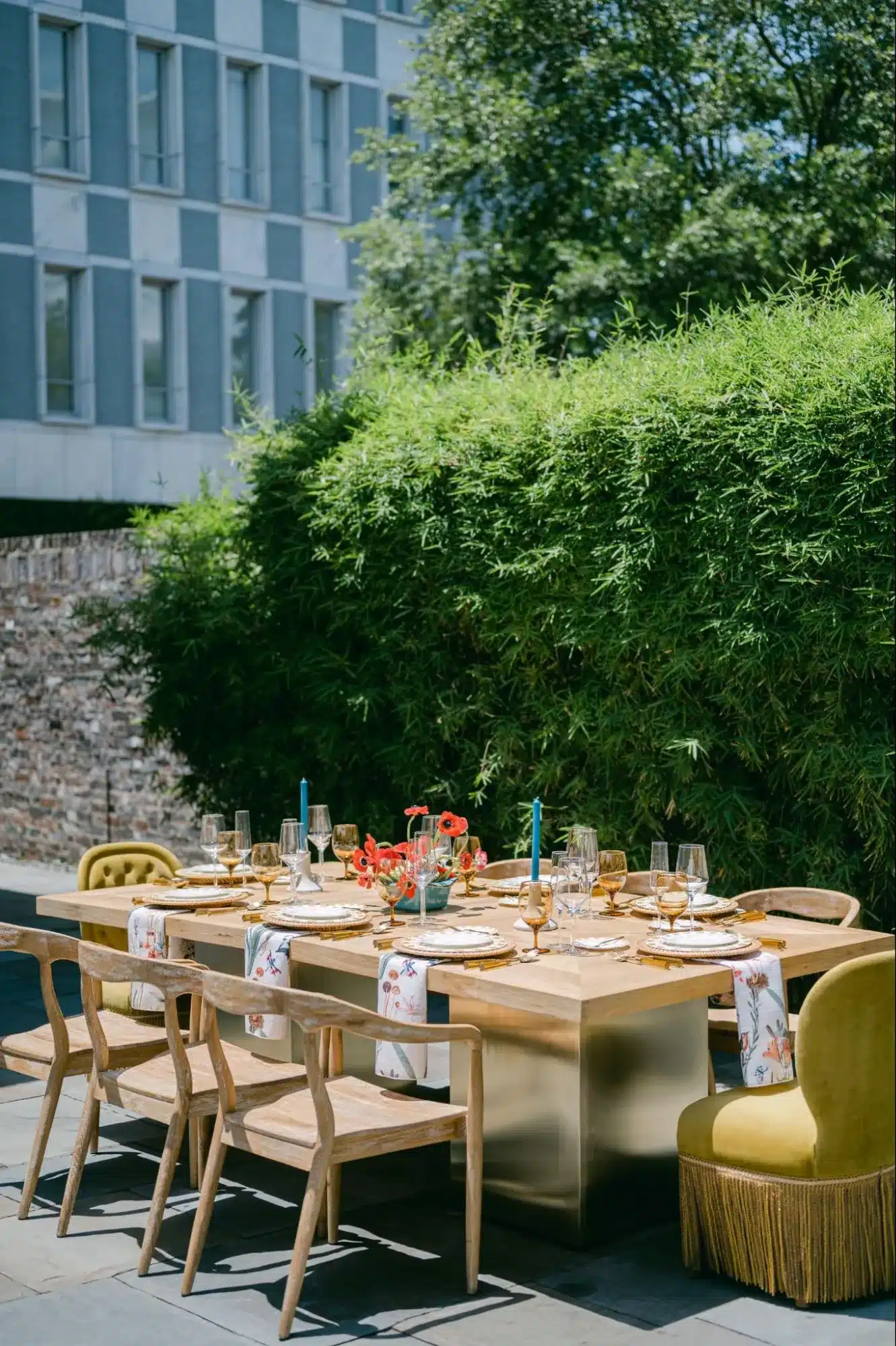 Outdoor brunch table with wood chairs, gold flatware, and vibrant floral accents against a lush hedge.