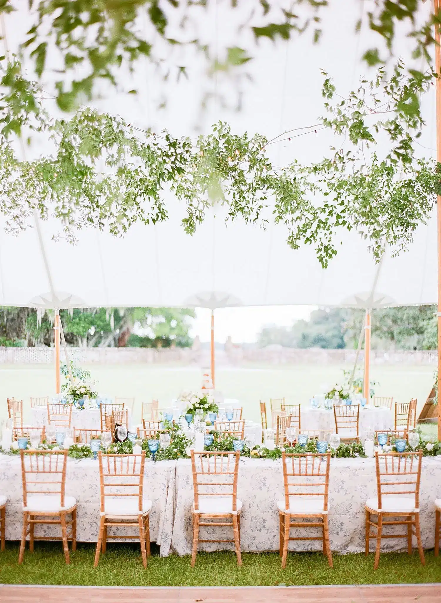 Elegant outdoor dining setup with decorated tables, floral arrangements, and wooden chairs under a white tent.