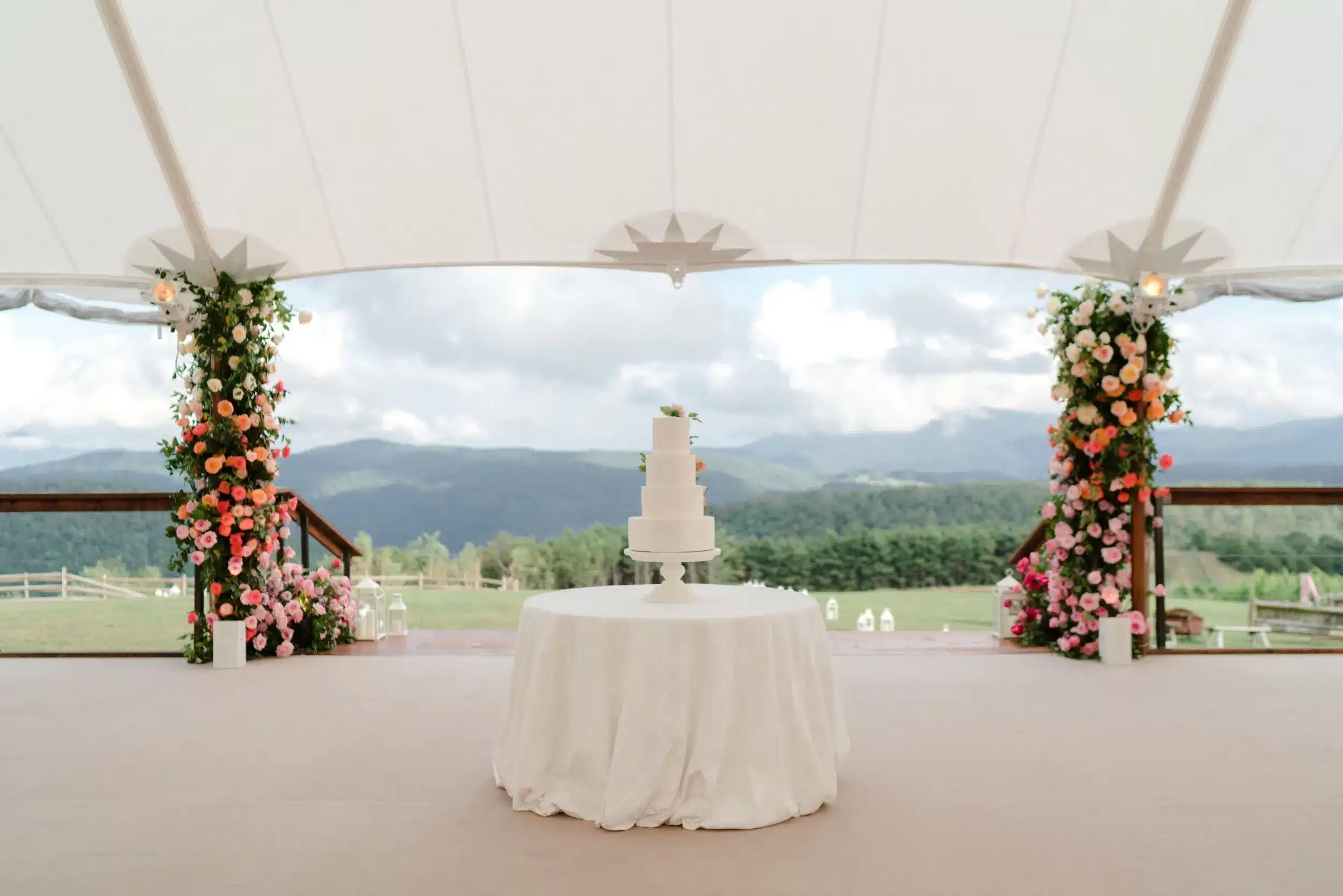 White tiered wedding cake on a round table beneath a tent, overlooking rolling hills and floral pillars.