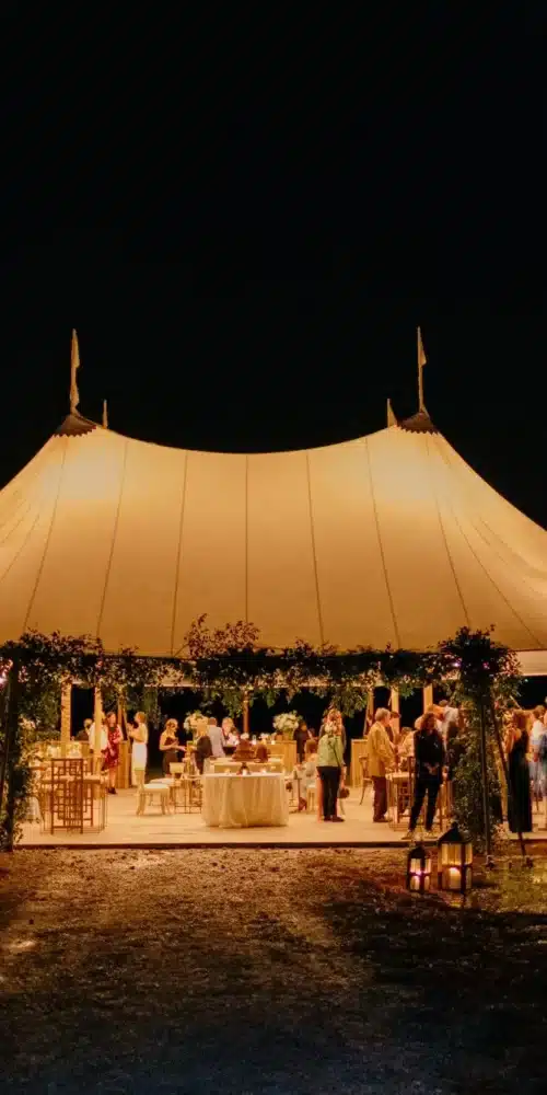 Illuminated sailcloth wedding tent at night with guests mingling and reception tables inside.