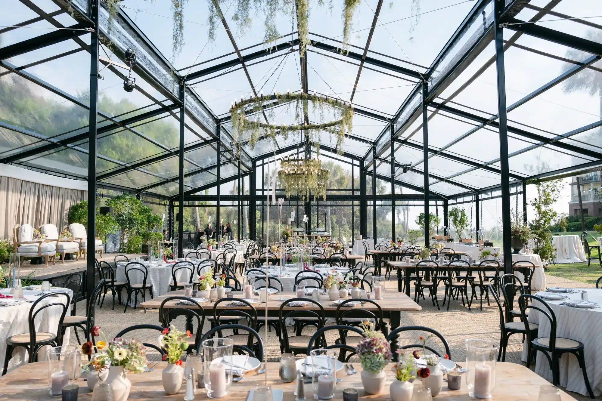 Open glass pavilion with widely spaced dining tables and hanging chandeliers. | Curated Events Open glass pavilion with widely spaced dining tables and hanging chandeliers.