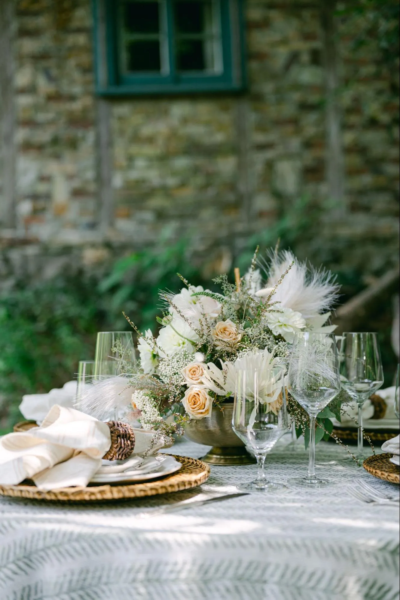 Elegant table setting with floral centerpiece featuring white and peach roses, surrounded by glassware and decorative napkins on a patterned tablecloth, against a rustic stone wall backdrop.