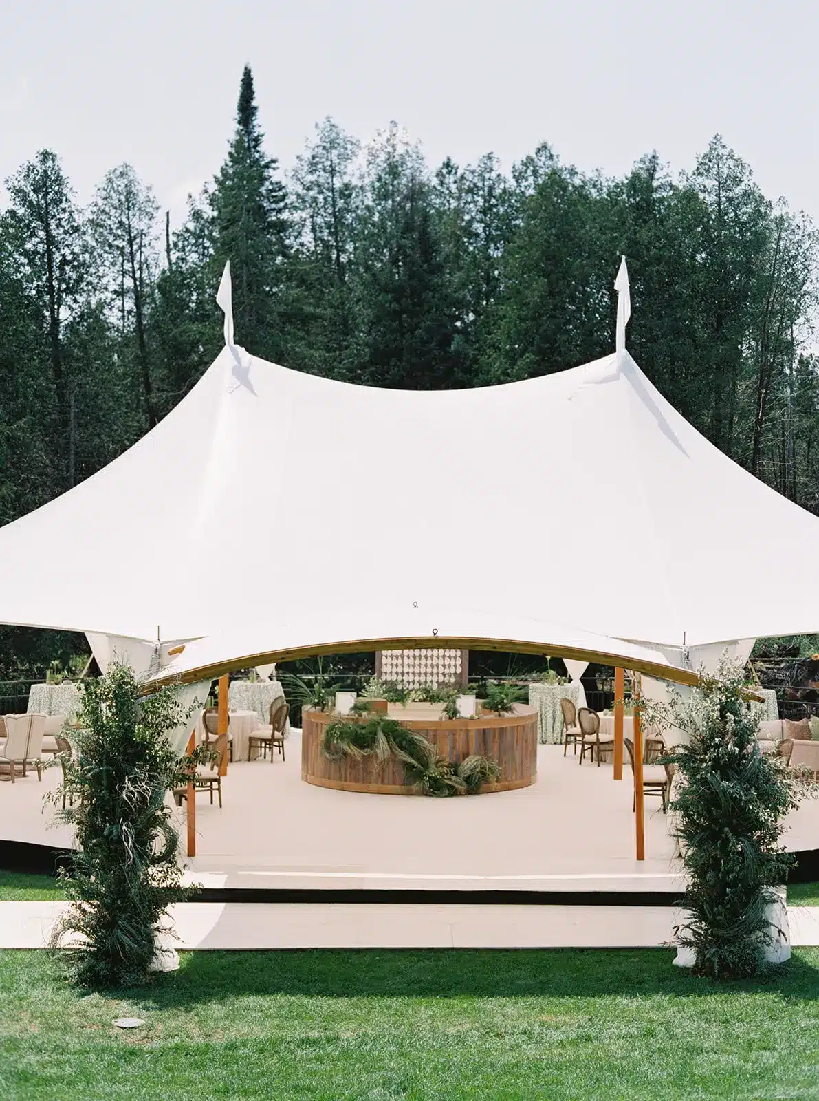 Open sailcloth tent with circular wood bar and greenery accents on a lawn.