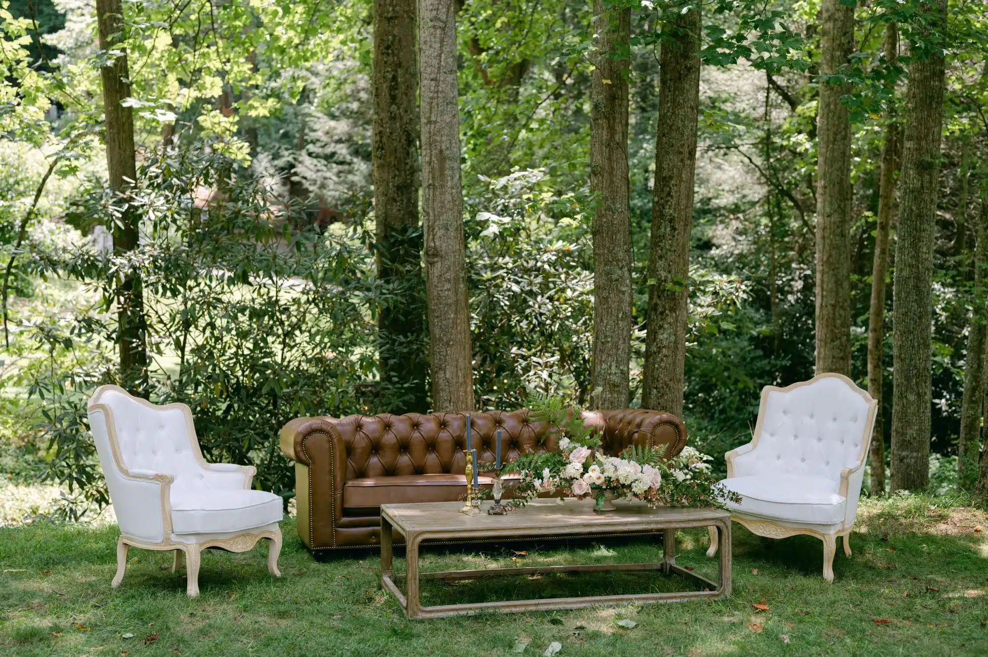 Chic outdoor lounge setup featuring a brown tufted leather sofa, white upholstered armchairs, and a rustic wooden coffee table adorned with floral arrangements and candles, ideal for an elevated bachelorette celebration.
