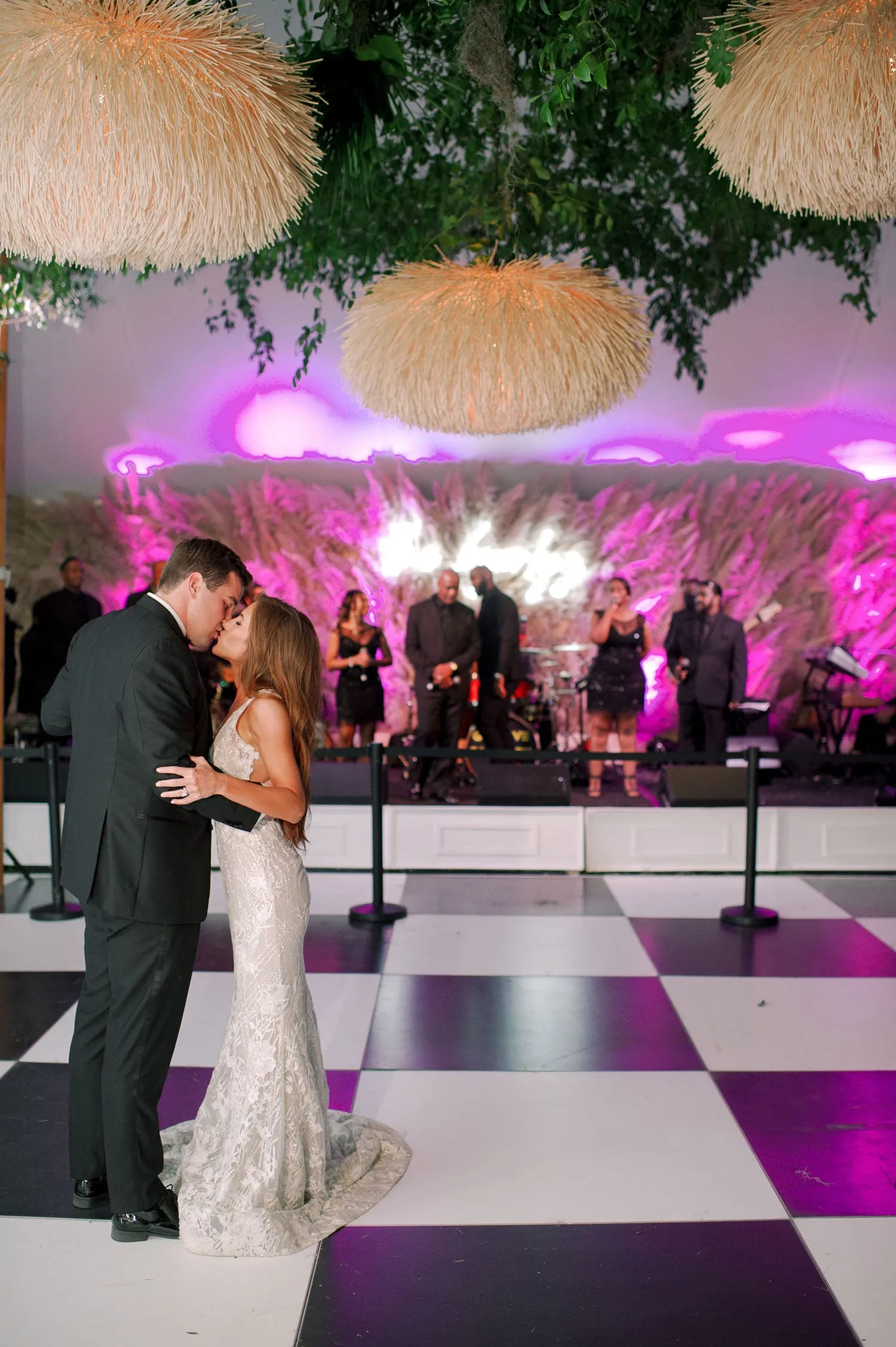 Couple sharing a romantic kiss on a dance floor, adorned with elegant hanging lights and a live band in the background, celebrating a special occasion.