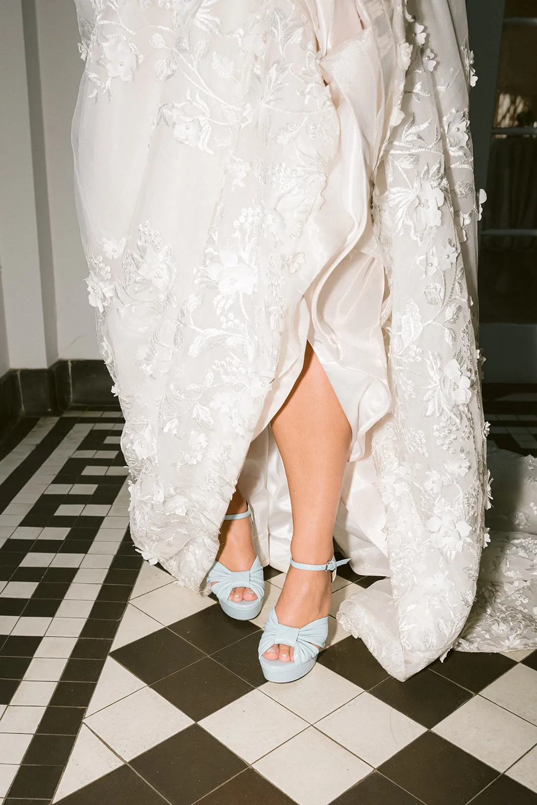 Bride's feet in blue platform shoes, partially visible under an intricately embroidered white wedding gown, standing on a black and white tiled floor.