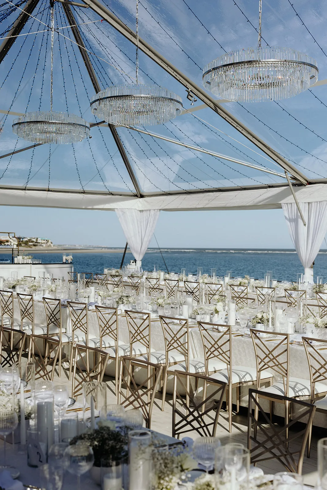 Coastal tent reception with chandeliers and long dining tables overlooking the water